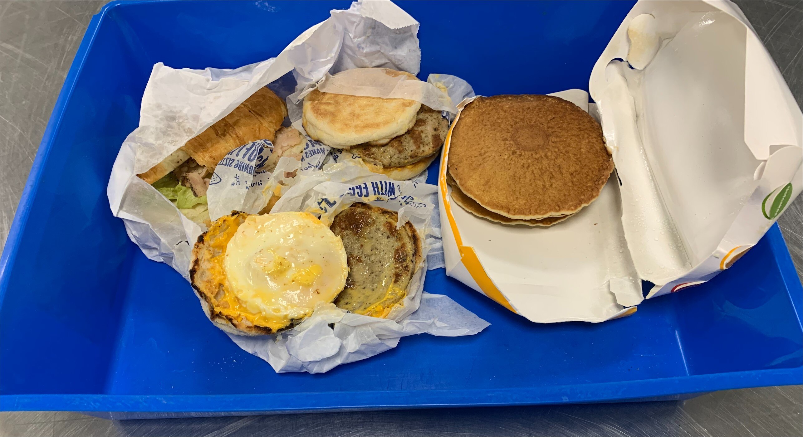 Two opened McDonald's mcmuffins, hot cakes and a croissant sit in opened wrapping on a blue plastic tub