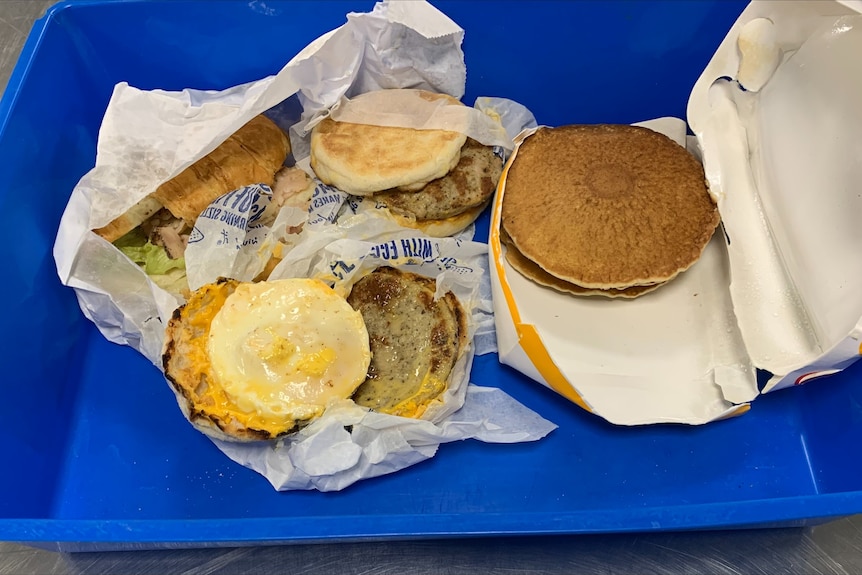 Two opened McDonald's mcmuffins, hot cakes and a croissant sit in opened wrapping on a blue plastic tub