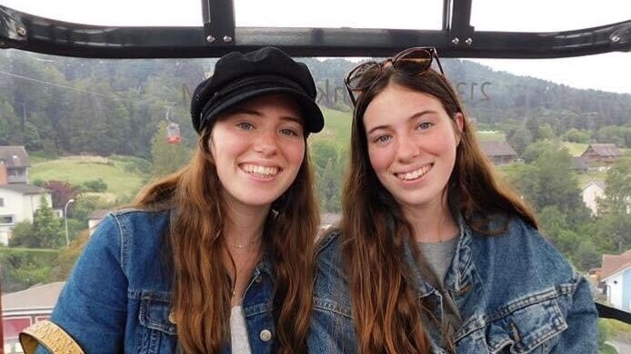 Two twin sisters sit in a carriage smiling at camera. Background is of green hills.