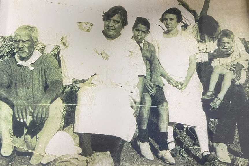 a black and white picture of a man sitting down, a woman holding a baby, and three young people and child