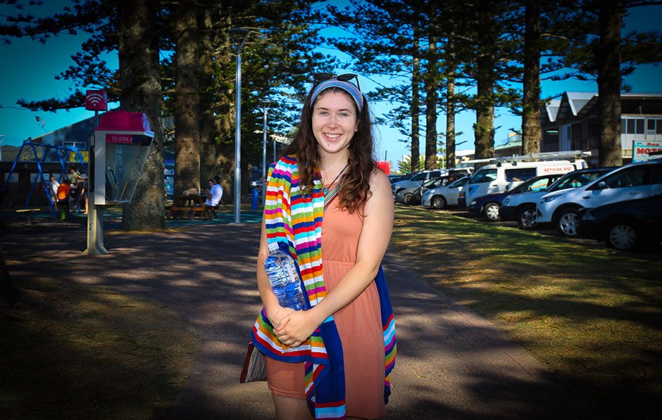 A young woman standing on a pathway with a striped towel over her shoulder, holding a bottle of water.