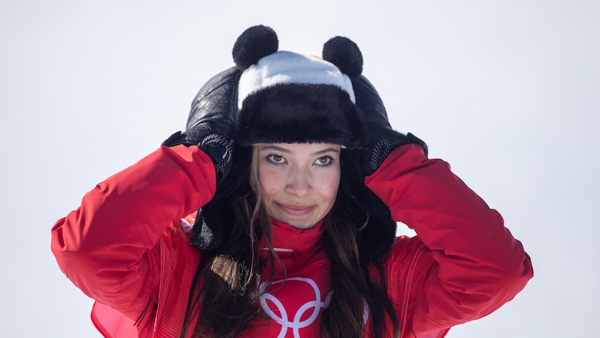 A girl in a red jacket and a hat with panda ears touches her head and smiles