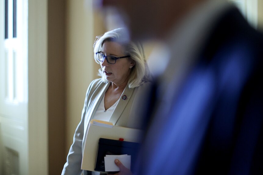 Liz Cheney, in a cream suit, clutches a stack of folders and notebooks as she exits a room. An unidentified man is out of focus 