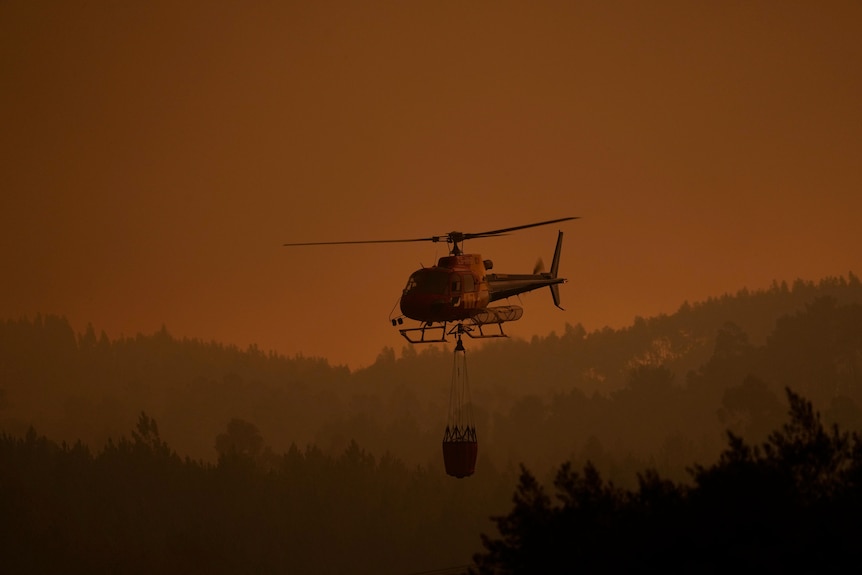 Helikopter strażacki leci nisko wśród gęstego dymu z pożaru lasu w wiosce w Portugalii.