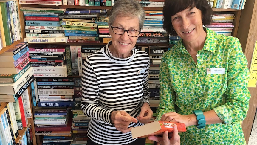 Volunteers Yvonne White and Paula Ryan stand in front of a massive bookshelf crammed with books.