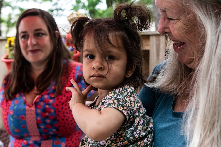 A toddler smiles at the camera as her grandmother smiles at her 