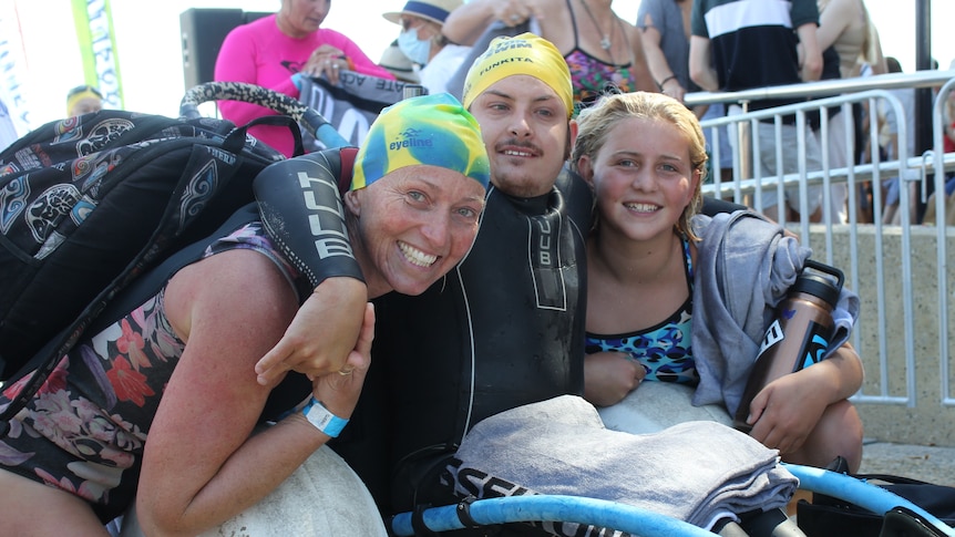 A man in wheelchair with a swimming cap, with two women also in swimming caps. Outside.