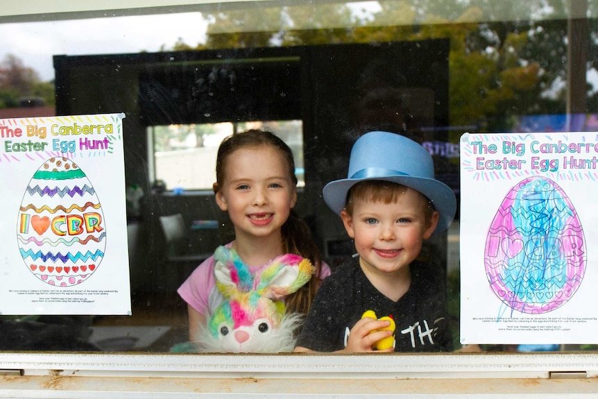 Two small children hold up their easter colouring in projects behind a window