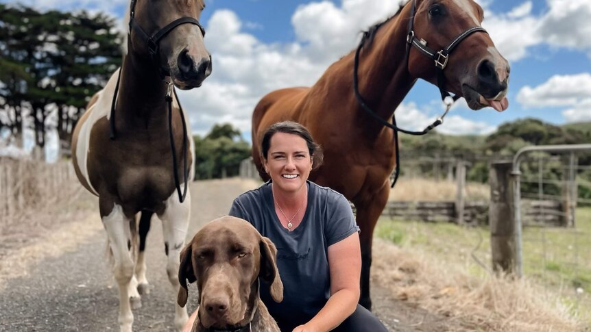 A woman smiling at the camera with her dog and two horses
