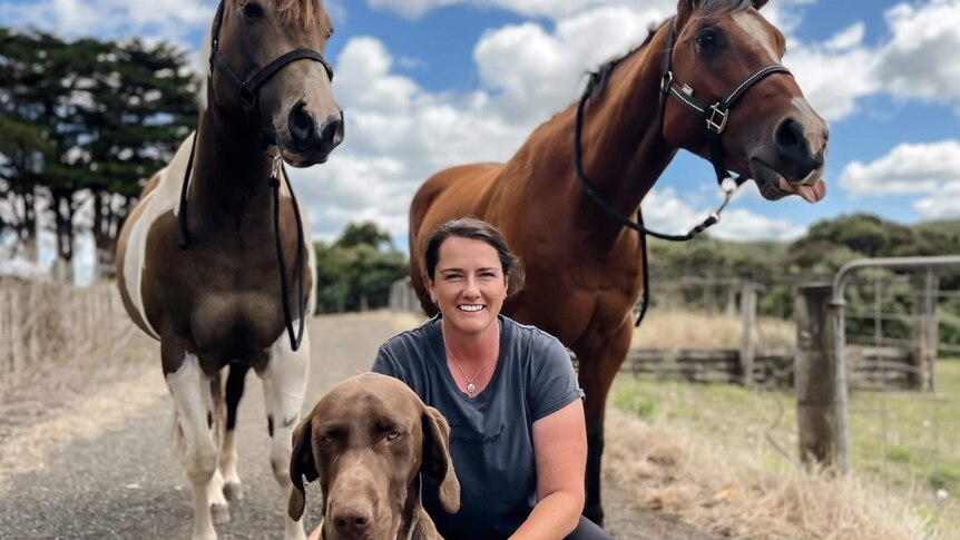A woman smiling at the camera with her dog and two horses