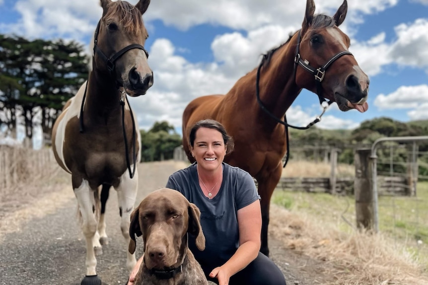 A woman smiling at the camera with her dog and two horses