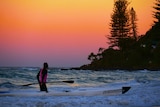 A Kayaker stands thigh-deep in water at the beach, next to their kayak, looking out at a beautiful pink sunrise.