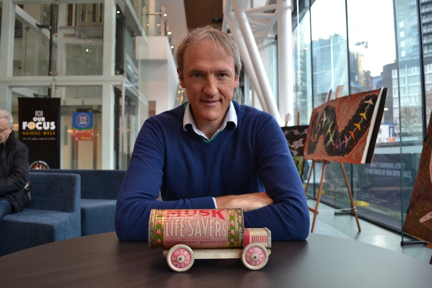 A man wearing a navy jumper over a shirt sits with his arms crossed, in front of an old toy truck labelled MUSK LIFESAVERS