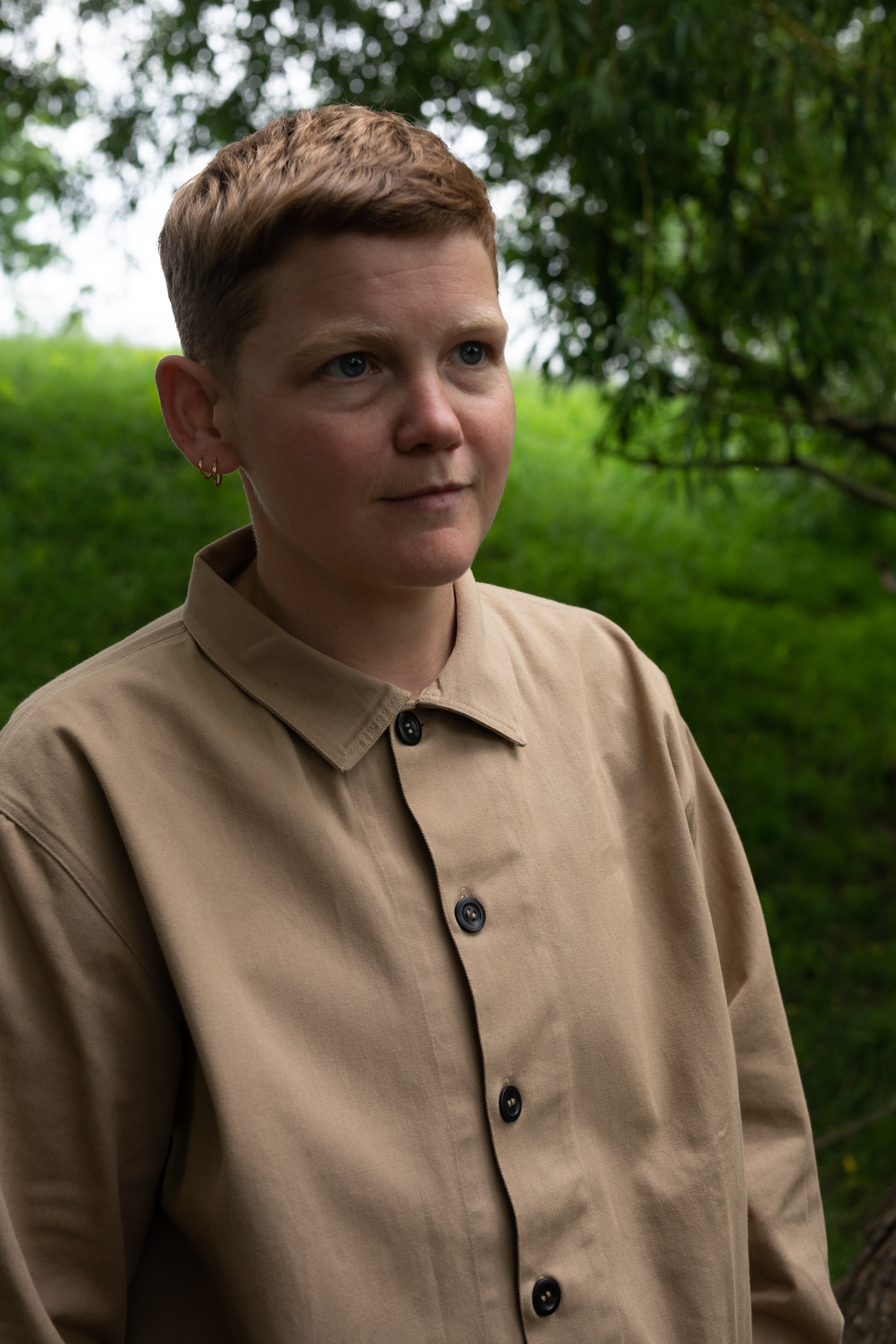 A white non-binary person with short sandy hair wears a long-sleeved khaki shirt and looks off-camera in a park.