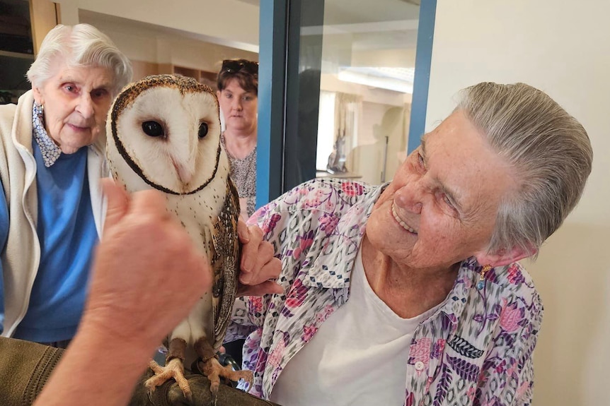 Two smiling older women look at a brown owl with a white face and chest.