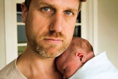 Close up image of man with cap with serious expression holding tiny, newborn baby wrapped in white blanket to his chest.