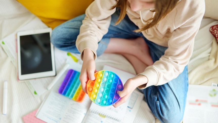Teenage girl playing with rainbow pop-it fidget toy while studying at home