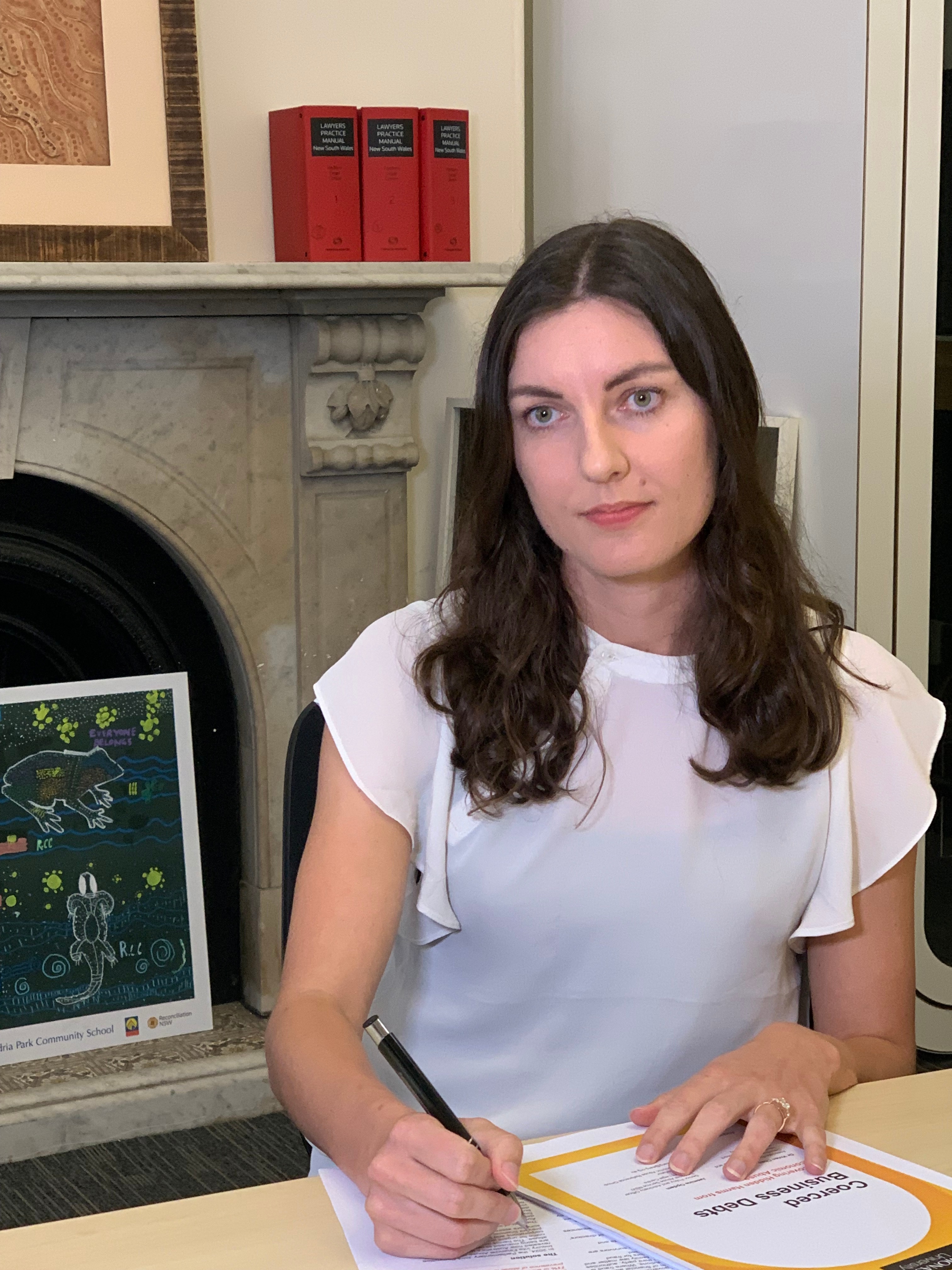 A lady in front of a fireplace sitting at a table with documents. She is wearing a white top and has long brown hair. 