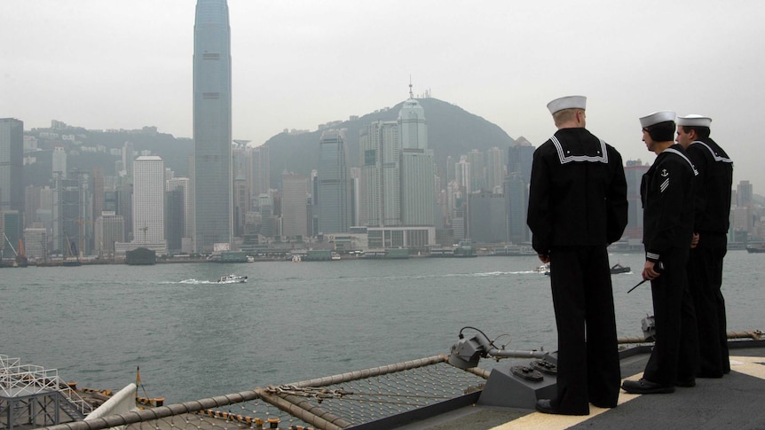 Three sailors on deck of a naval ship look out to the Hong Kong cityscape.