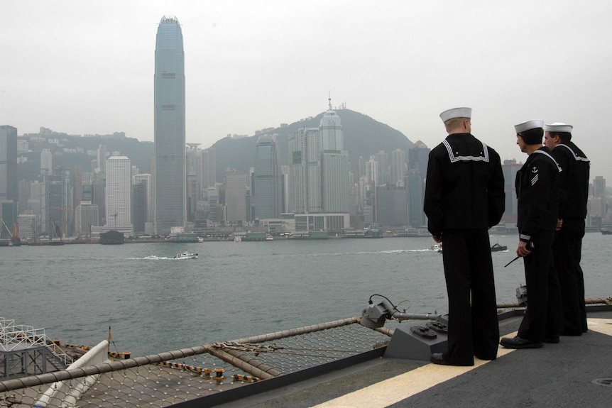Three sailors on deck of a naval ship look out to the Hong Kong cityscape.