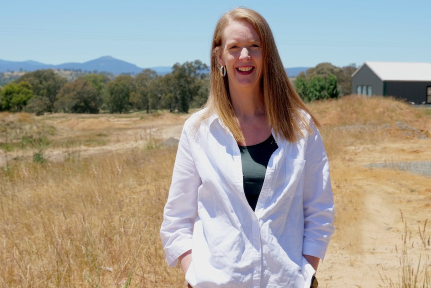 A woman standing in a paddock with a cabin behind her.