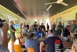 A large group of Menindee locals gathered under a shaded building space, with one man speaking to the assembled group