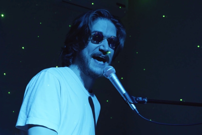 Bo performs at a piano while wearing sunglasses and casting a green light around a dark room. 
