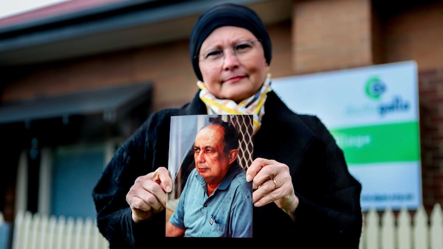 Woman in black coat and scarf wearing glasses stands outside aged care home holding photo of her father