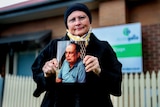 Woman in black coat and scarf wearing glasses stands outside aged care home holding photo of her father