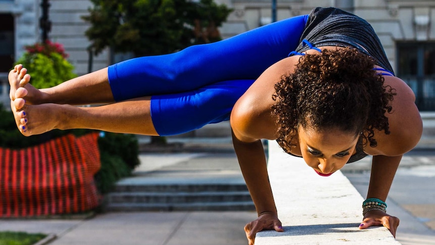 A young woman performs a yoga pose, balancing on a low wall with her hands, with both her legs extended out to one side.