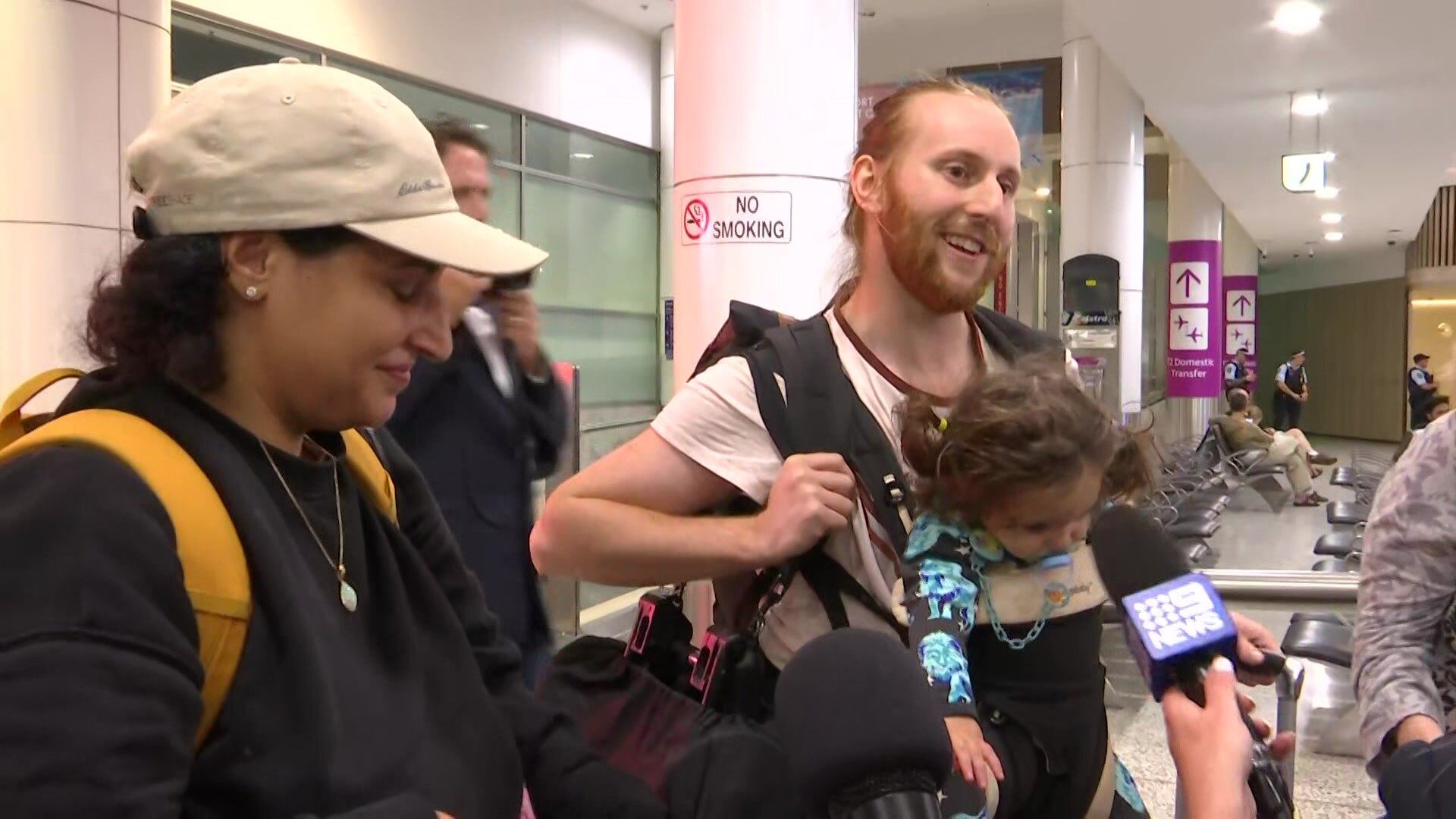 a man named Michael Landis with a young child strapped to his chest stands next to his wife at Sydney airport