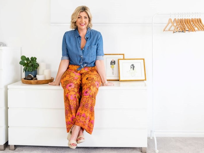 Woman who is 40, blonde hair, sitting on a cabinet with bright orange trousers and blue shirt. She is smiling.