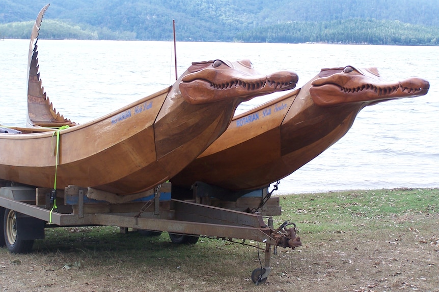 Two handmade wooden canoes in shape of crocodiles sitting side by side on a trailer beside Lake Tinaroo