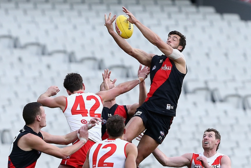 An AFL player in black and red catches a yellow football above opponents wearing white and red in empty stadium.