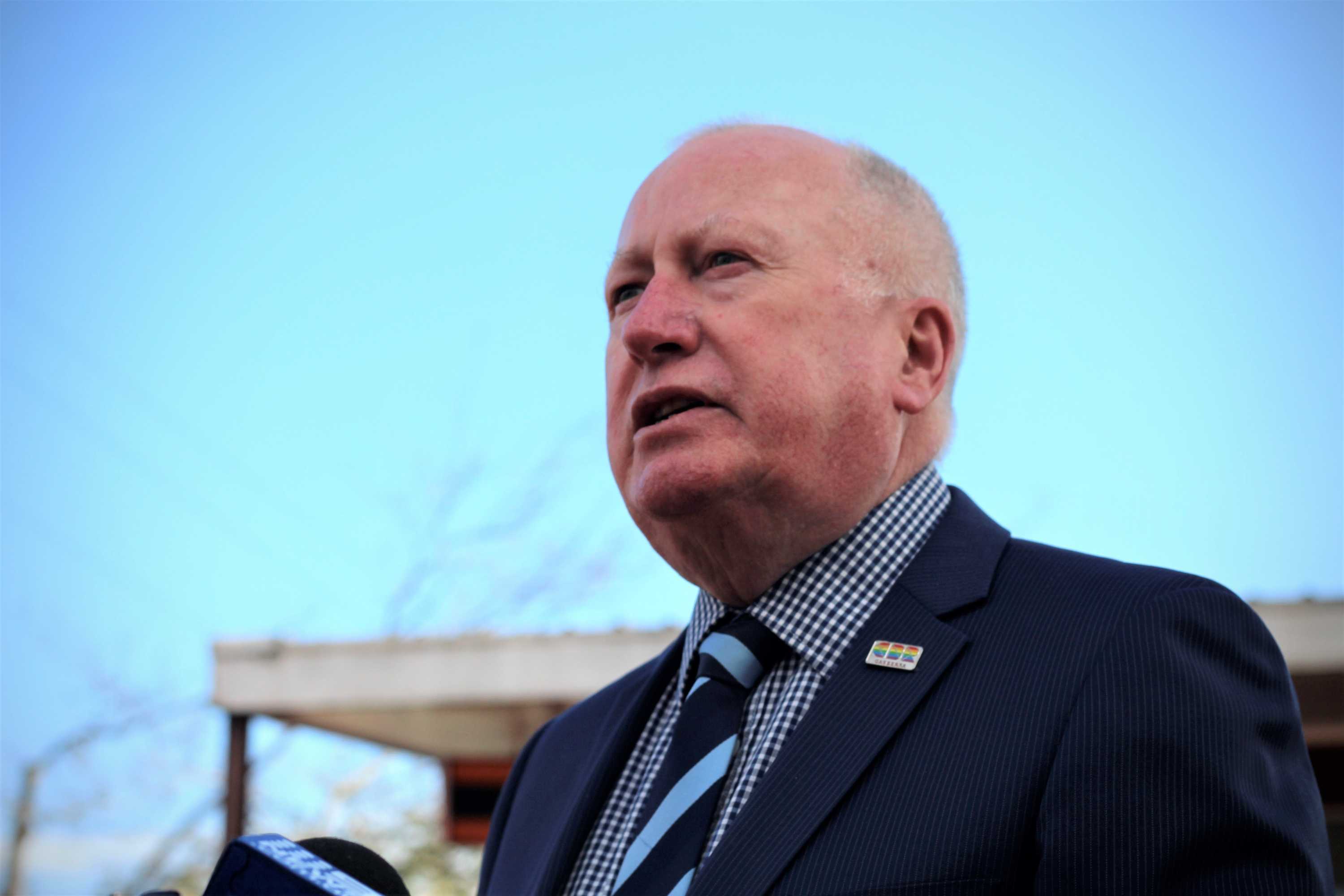 A man stares past the camera at a press conference wearing a suit and backdropped by a blue sky.