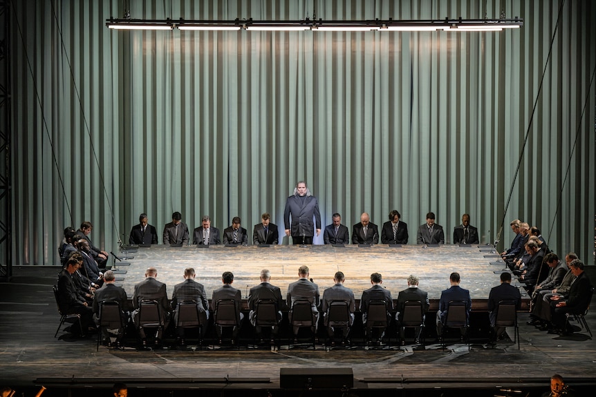 Forty men in suits sit around a large table set on stage against a drab green backdrop, with one standing in the centre.