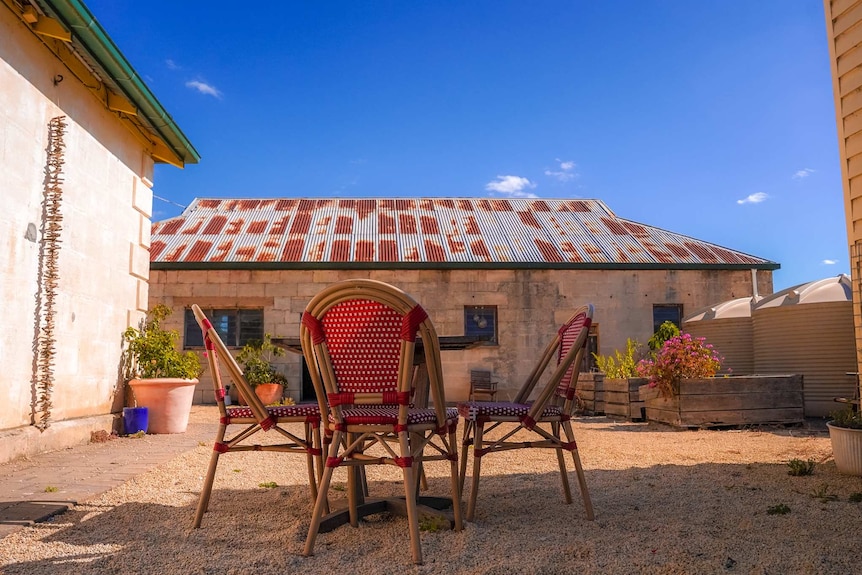 Four red and wood chairs on gravel in an outdoor terrace next to a stone cottage with a rusted roof on a sunny day.