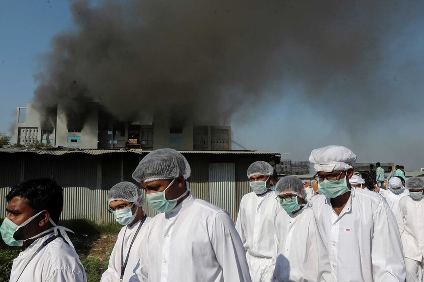 Workers in face masks, white lab gowns and hair nets walk in front of a burning building.