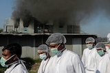 Workers in face masks, white lab gowns and hair nets walk in front of a burning building.