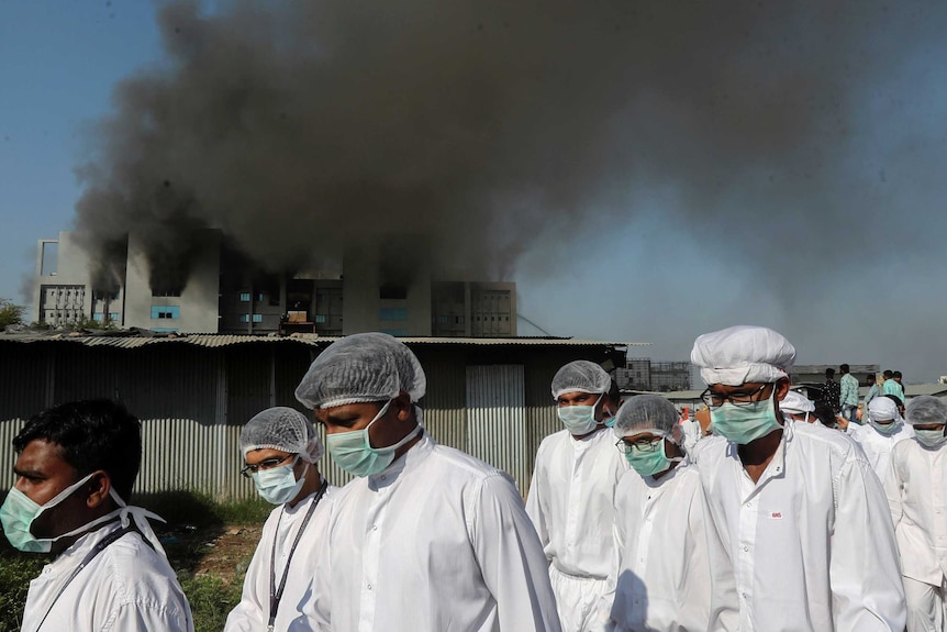 Workers in face masks, white lab gowns and hair nets walk in front of a burning building.