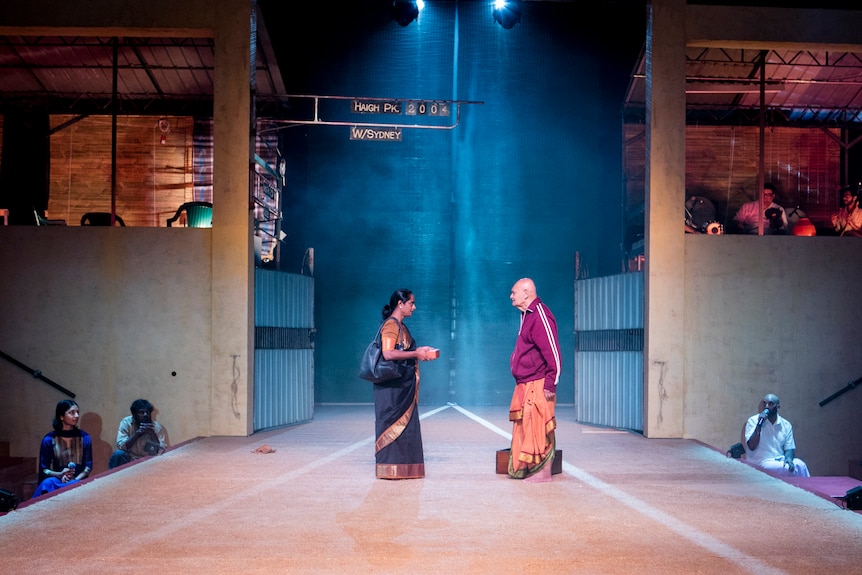 On a stage stand a Sri Lankan woman in a black sari speaking to a Sri Lankan man in a red adidas jacket and orange sarong