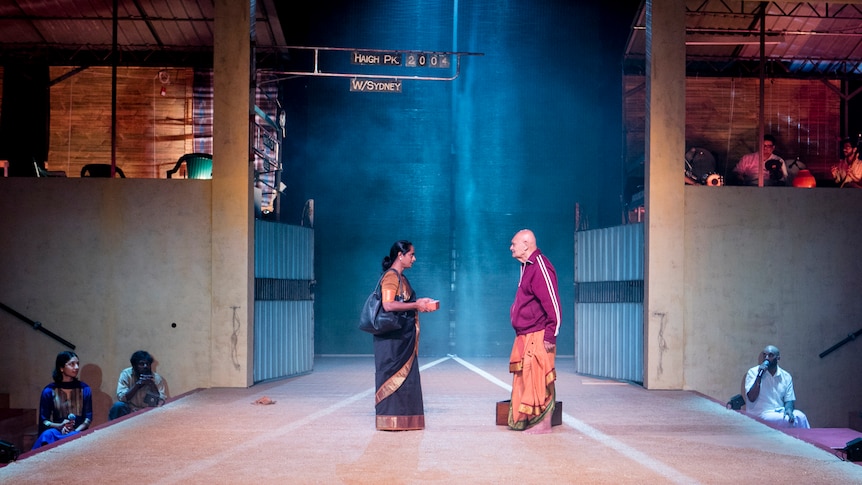 On a stage stand a Sri Lankan woman in a black sari speaking to a Sri Lankan man in a red adidas jacket and orange sarong
