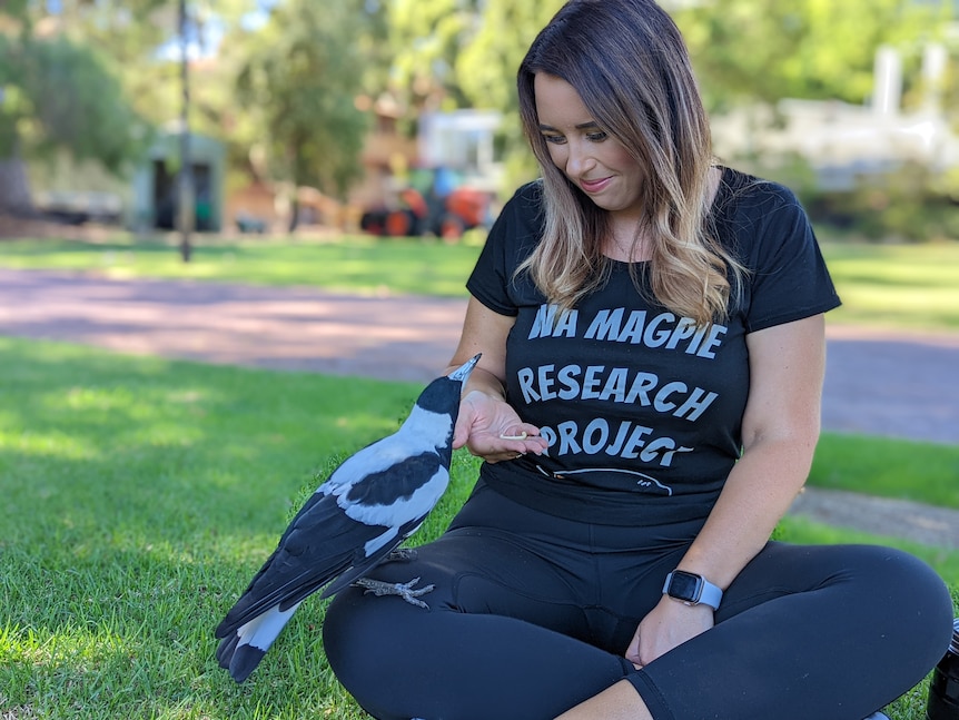 Woman with brown hair and black clothes sits on grass smiling at magpie on her lap