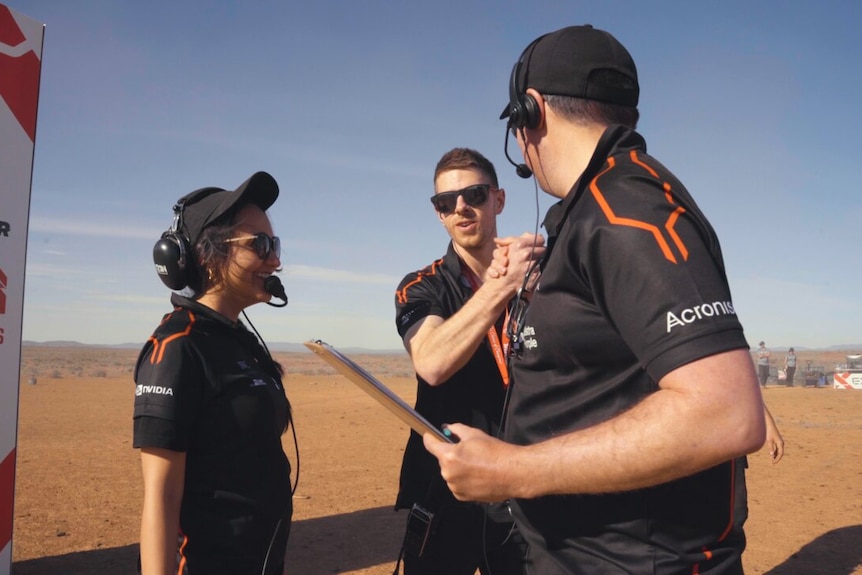 Three people in black polo shirts with headsets stand facing each other in a desert setting.