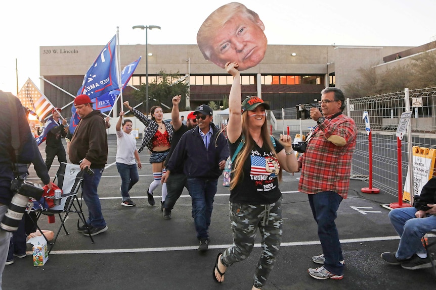 People holding Trump face posters and flags protesting in a car park at sunset