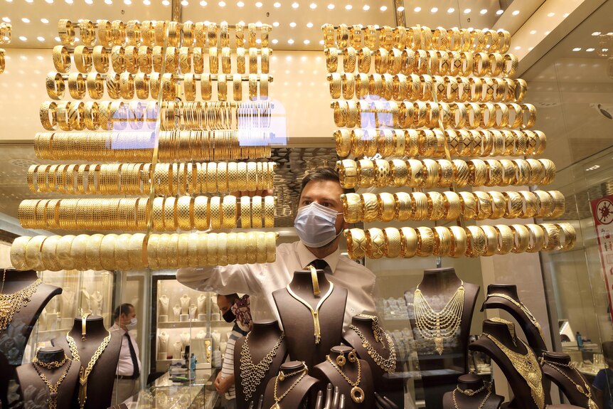 Un bijutier de aur purtând o mască de protecție aranjează brățări de aur în timp ce alții vorbesc cu clienții.