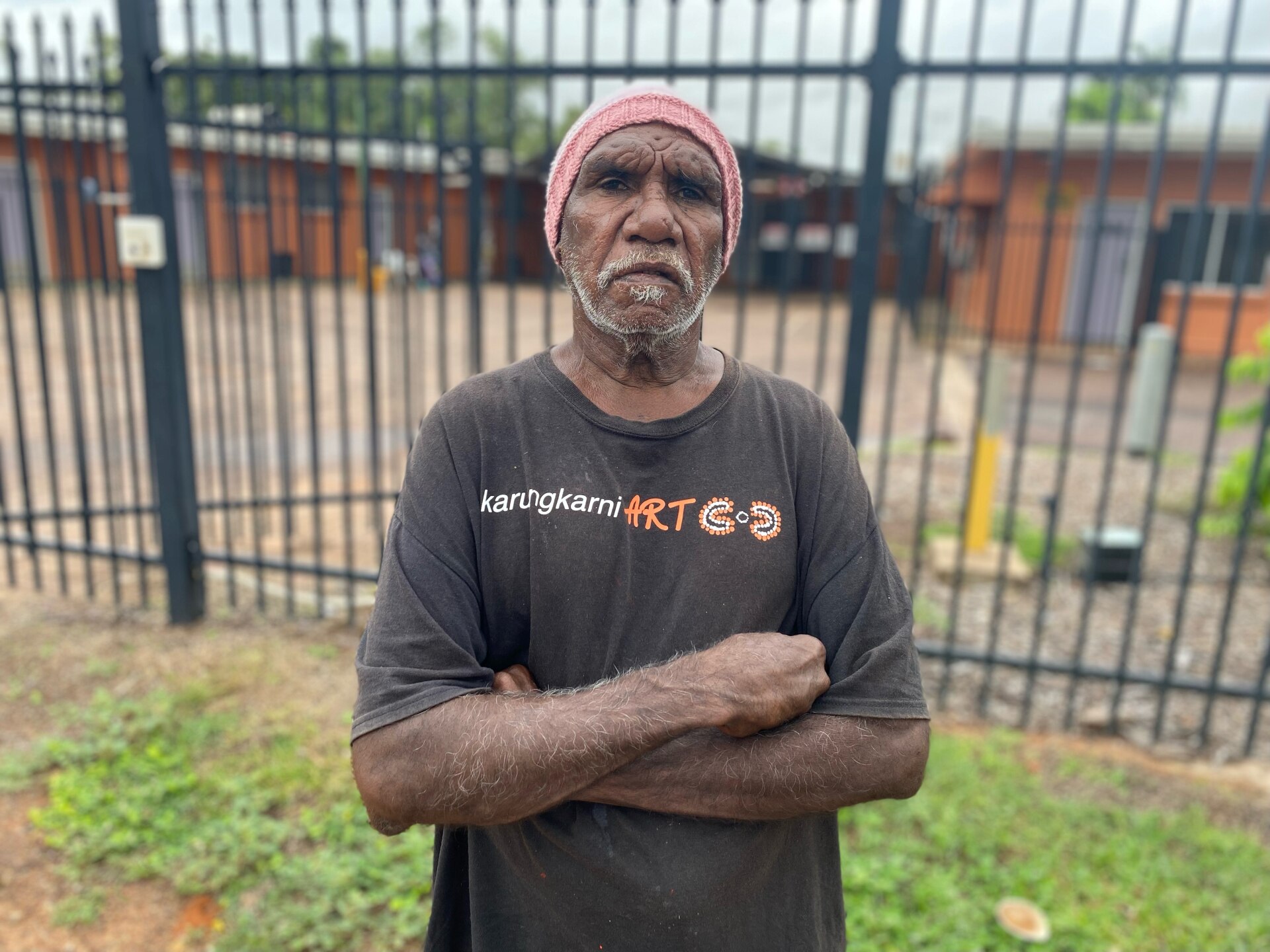 A man with his arms folded and looking serious, in front of a fence around a single-level orange building. 