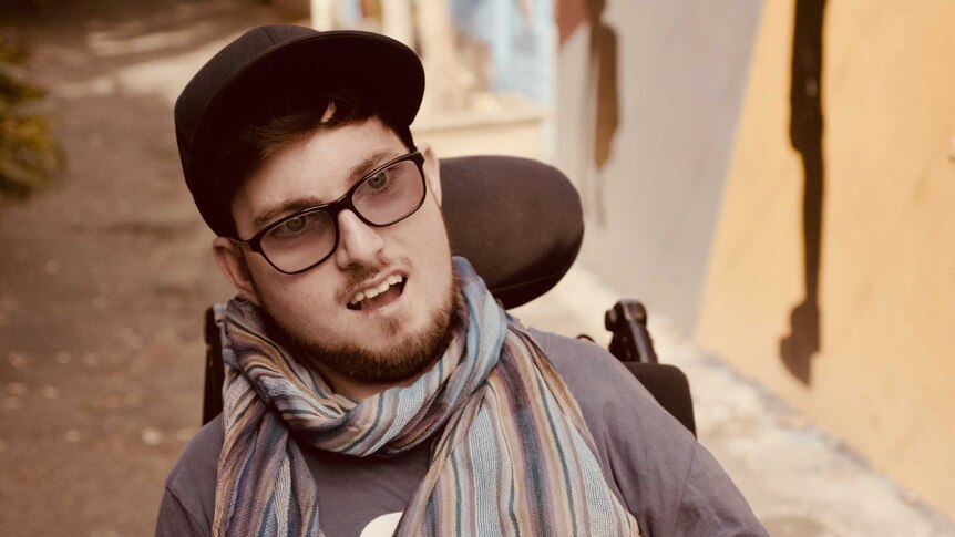 A portrait of a man in a hat and glasses, wearing a scarf, sitting in a wheelchair.