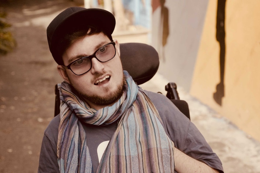 A portrait of a man in a hat and glasses, wearing a scarf, sitting in a wheelchair.
