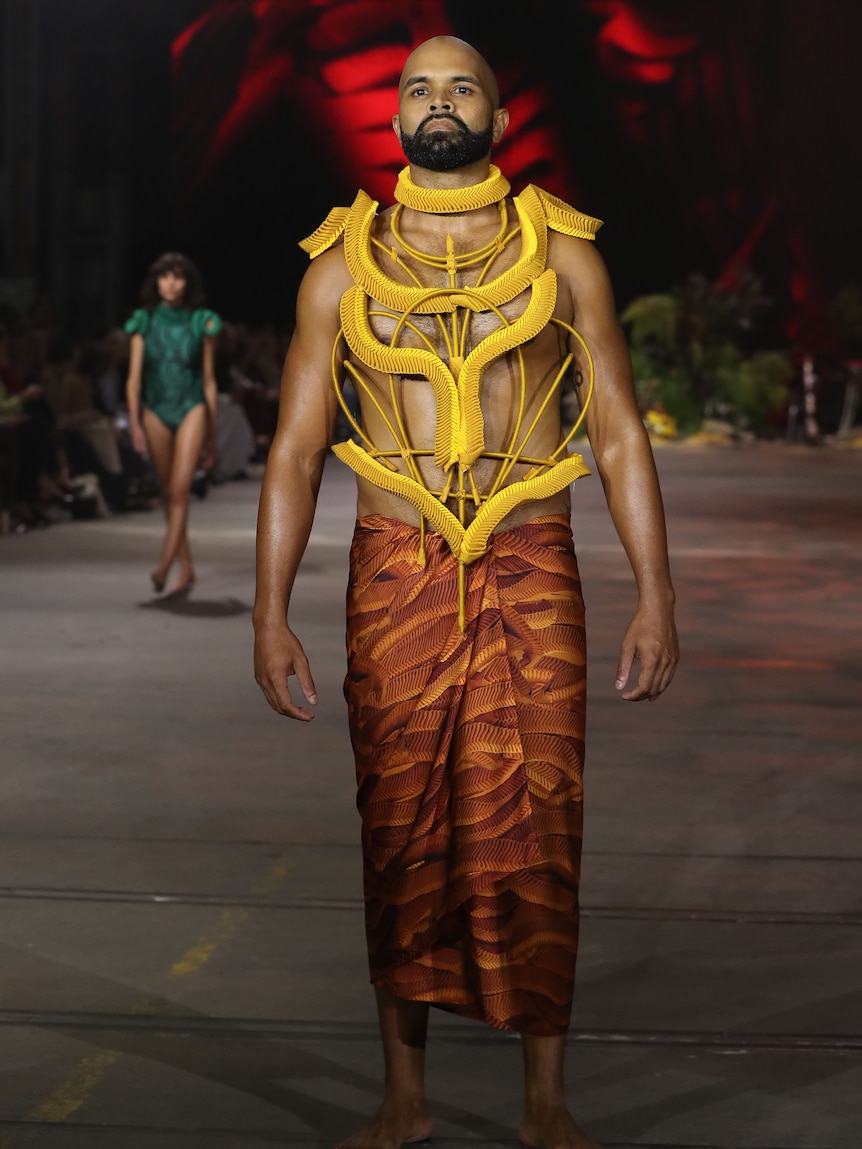 Indigenous man wears vibrant yellow sculptured garment made with weaving technique on fashion runway.